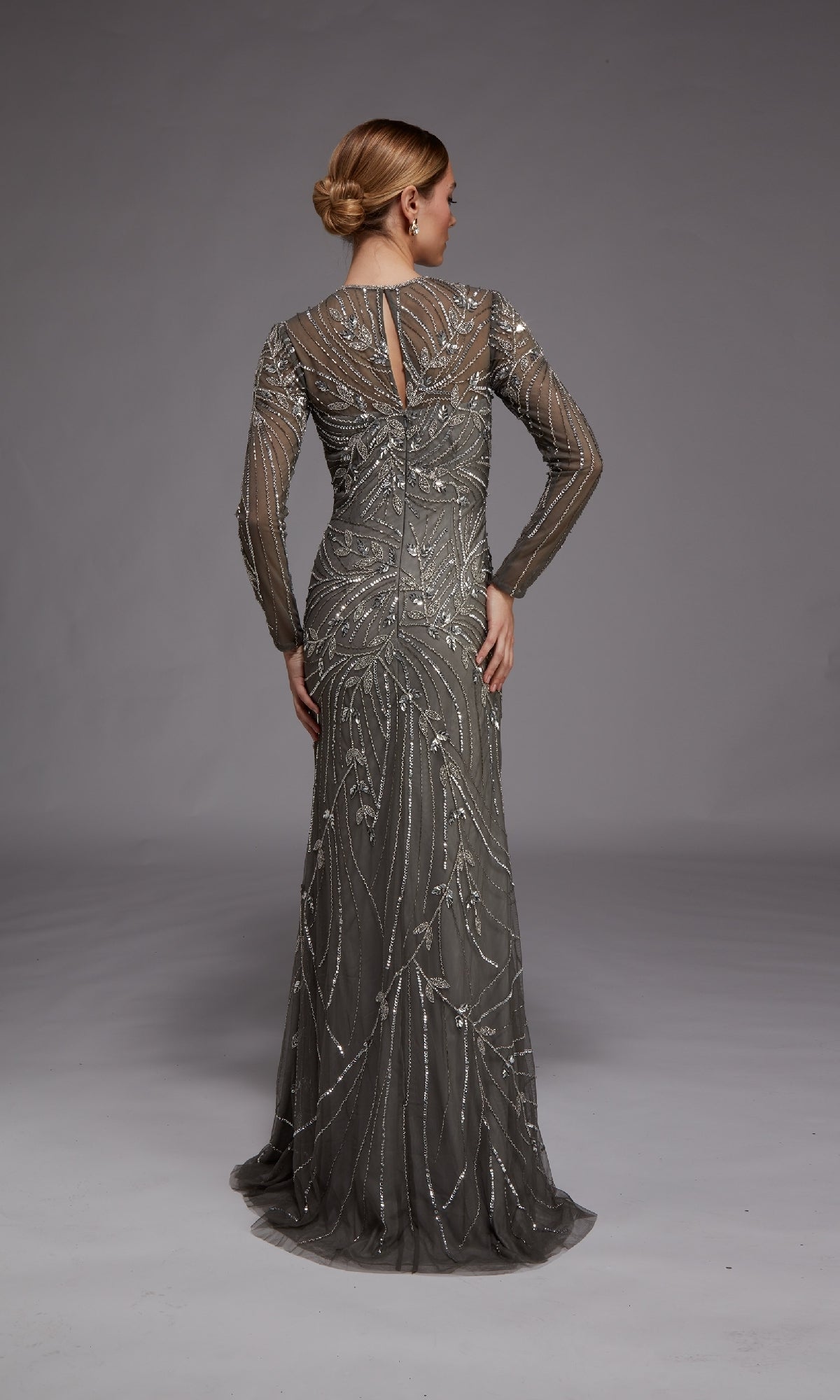 A woman faces away, wearing the Alyce 27801 long formal dress—a fitted grey evening gown with sheer sleeves and intricate silver bead embellishments—set against a plain grey background.